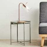 Grey iron side table with a sleek, modern design featuring a glass top and a geometric base, accompanied by a stylish lamp with a curved neck and a warm light, positioned next to a grey upholstered sofa on a light-colored floor