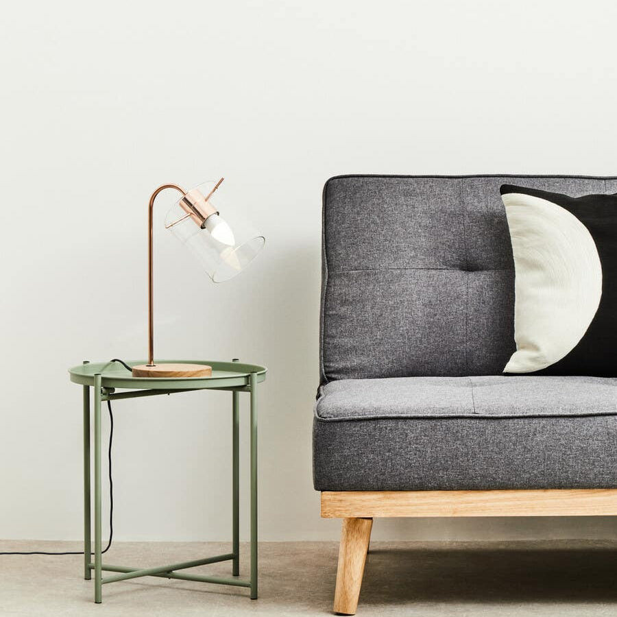 A green iron side table with a minimalist design featuring a square top and slender legs, positioned next to a modern gray sofa with tufted upholstery and a round black and white pillow. A stylish lamp with a curved neck and white shade rests on the table, set against a light-colored wall and neutral floor