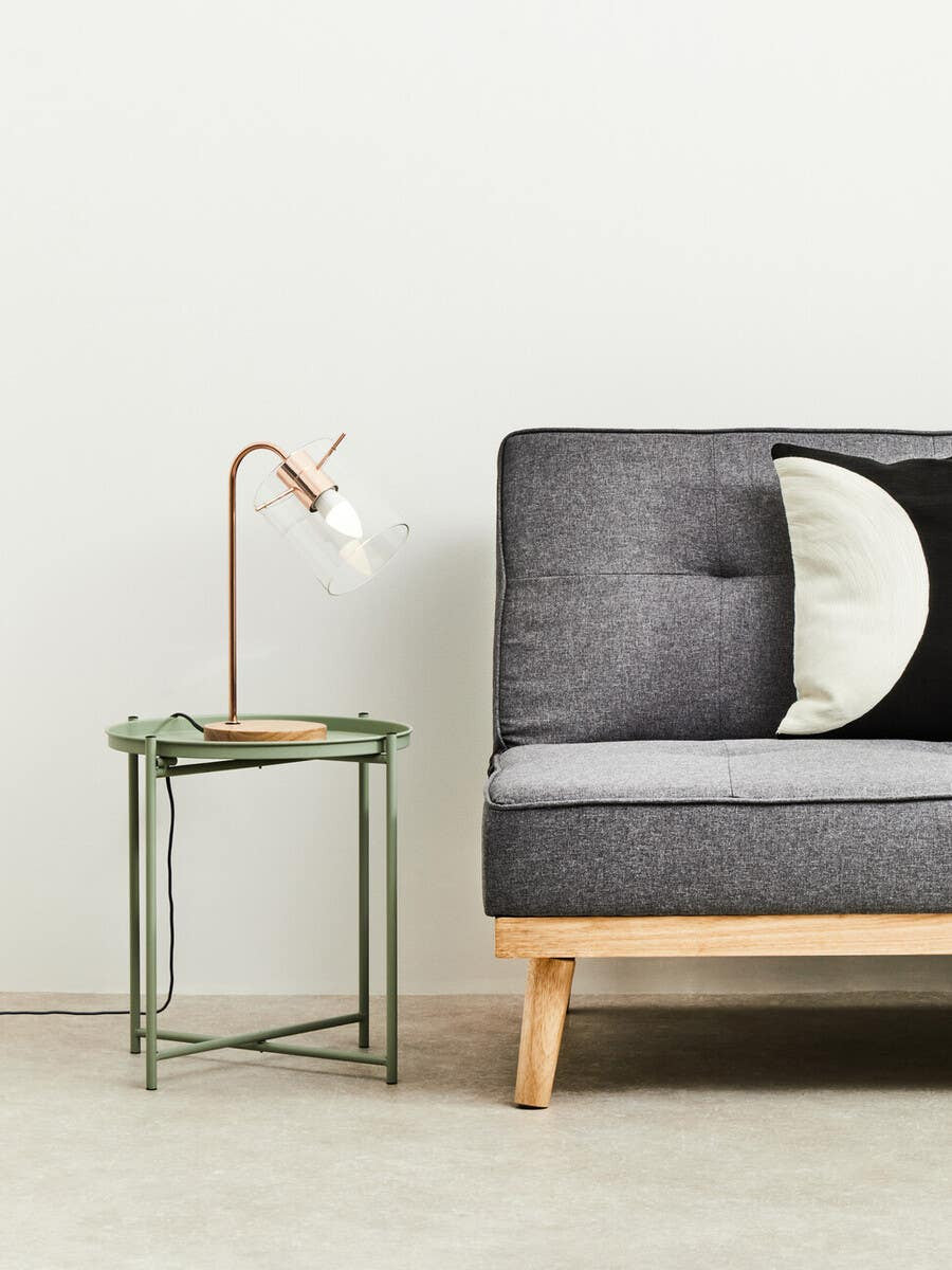 A green iron side table with a minimalist design featuring a square top and slender legs, positioned next to a modern gray sofa with tufted upholstery and a round black and white pillow. A stylish lamp with a curved neck and white shade rests on the table, set against a light-colored wall and neutral floor