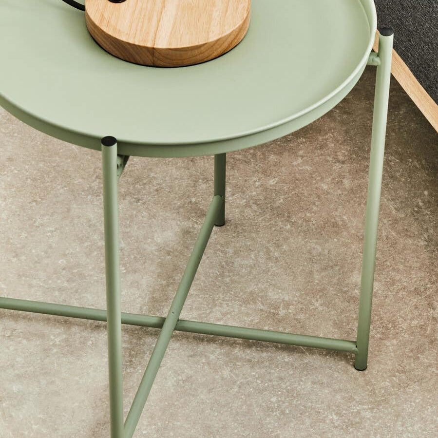 A green iron side table with a round tray top and slender legs, positioned on a light-colored carpet. A wooden lamp base is placed on the table, complementing the minimalist design
