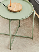 A green iron side table with a round tray top and slender legs, positioned on a light-colored carpet. A wooden lamp base is placed on the table, complementing the minimalist design