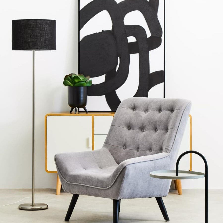 A modern living space featuring a grey and black hanging top side table next to a stylish grey armchair with button detailing. A tall black floor lamp stands nearby, and a potted plant adds a touch of greenery. Behind the chair, an abstract black and white artwork is displayed on the wall, complementing the contemporary decor