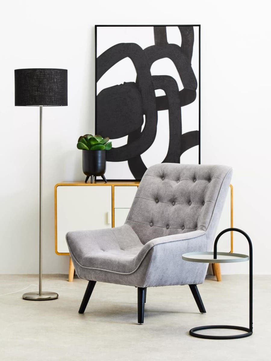 A modern living space featuring a grey and black hanging top side table next to a stylish grey armchair with button detailing. A tall black floor lamp stands nearby, and a potted plant adds a touch of greenery. Behind the chair, an abstract black and white artwork is displayed on the wall, complementing the contemporary decor