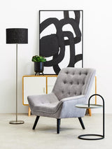 A modern living space featuring a grey and black hanging top side table next to a stylish grey armchair with button detailing. A tall black floor lamp stands nearby, and a potted plant adds a touch of greenery. Behind the chair, an abstract black and white artwork is displayed on the wall, complementing the contemporary decor
