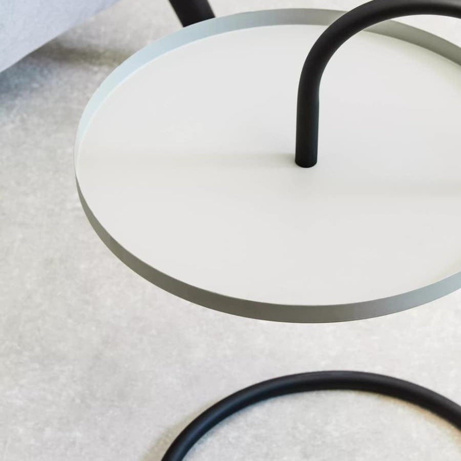 A round, grey and black side table featuring a sleek, minimalist design. The tabletop is circular and grey, supported by a black, curved metal frame that creates a hanging effect. The surface is smooth and modern, set against a light-colored background