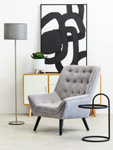 A modern living space featuring a Trosa Black Hanging Top Side Table next to a light gray upholstered armchair with button detailing, a sleek floor lamp, and a minimalist sideboard with a black and white abstract art piece on the wall. The side table has a circular top and a slender, curved base