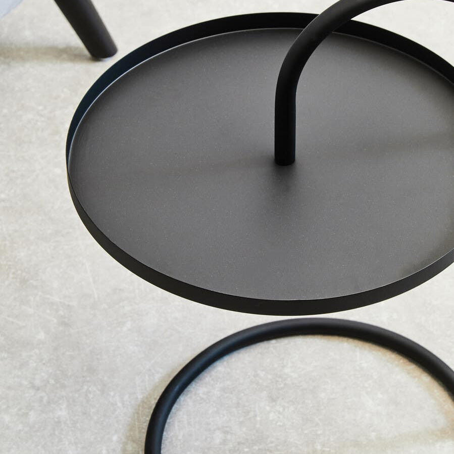 Black hanging top side table with a circular surface and a curved support leg, set against a light-colored carpet. The design features a minimalist aesthetic with clean lines and a modern look