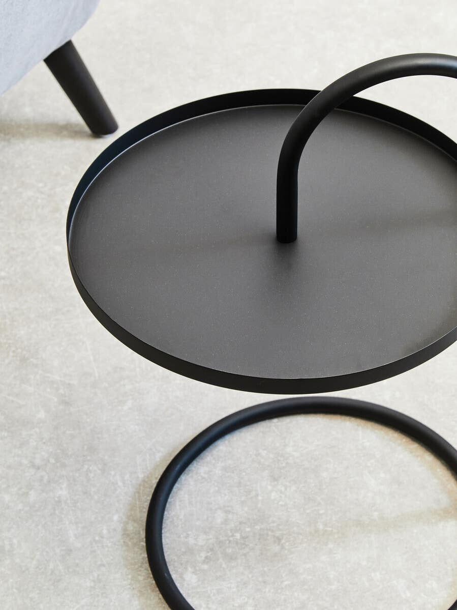 Black hanging top side table with a circular surface and a curved support leg, set against a light-colored carpet. The design features a minimalist aesthetic with clean lines and a modern look