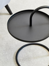 Black hanging top side table with a circular surface and a curved support leg, set against a light-colored carpet. The design features a minimalist aesthetic with clean lines and a modern look
