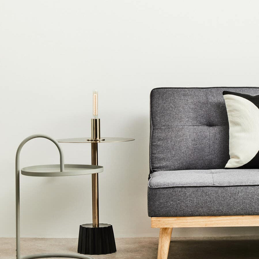 Grey hanging top side table with a curved metal frame, positioned next to a modern grey sofa adorned with a decorative pillow. A small black broom is placed beside the table on a light wooden floor. The background features a plain white wall