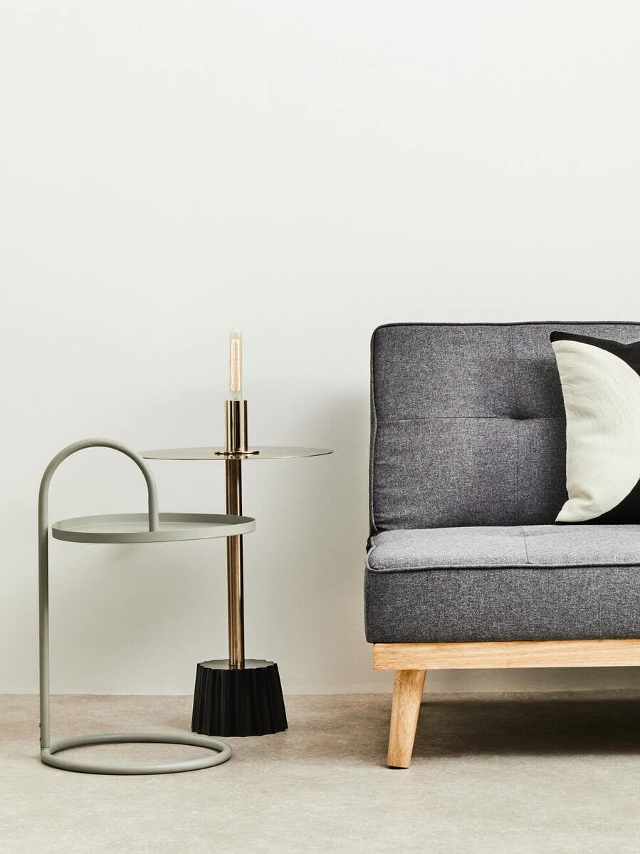Grey hanging top side table with a curved metal frame, positioned next to a modern grey sofa adorned with a decorative pillow. A small black broom is placed beside the table on a light wooden floor. The background features a plain white wall