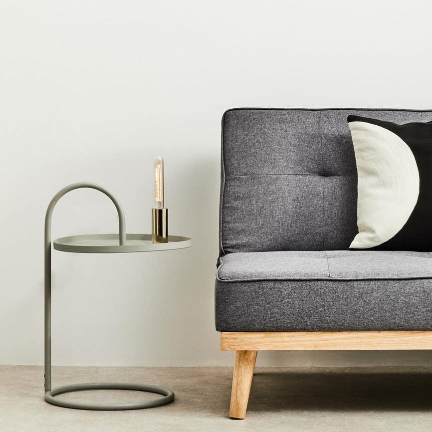 Grey hanging top side table with a circular base and a sleek, modern design, positioned next to a dark grey sofa with tufted upholstery and a decorative black and white pillow. A candle is placed on the table, enhancing the contemporary aesthetic of the scene. The background features a light-colored wall and a neutral floor