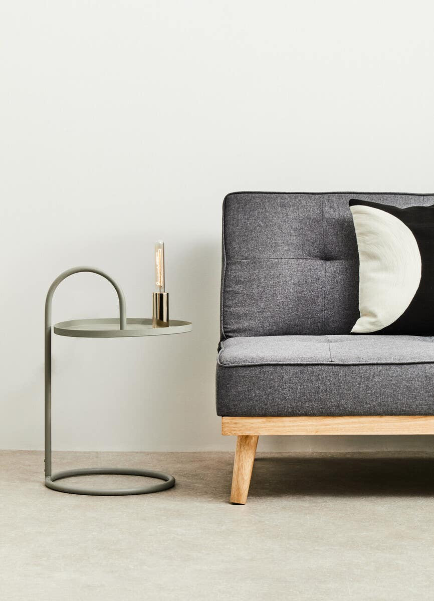 Grey hanging top side table with a circular base and a sleek, modern design, positioned next to a dark grey sofa with tufted upholstery and a decorative black and white pillow. A candle is placed on the table, enhancing the contemporary aesthetic of the scene. The background features a light-colored wall and a neutral floor