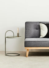 Grey hanging top side table with a circular base and a sleek, modern design, positioned next to a dark grey sofa with tufted upholstery and a decorative black and white pillow. A candle is placed on the table, enhancing the contemporary aesthetic of the scene. The background features a light-colored wall and a neutral floor