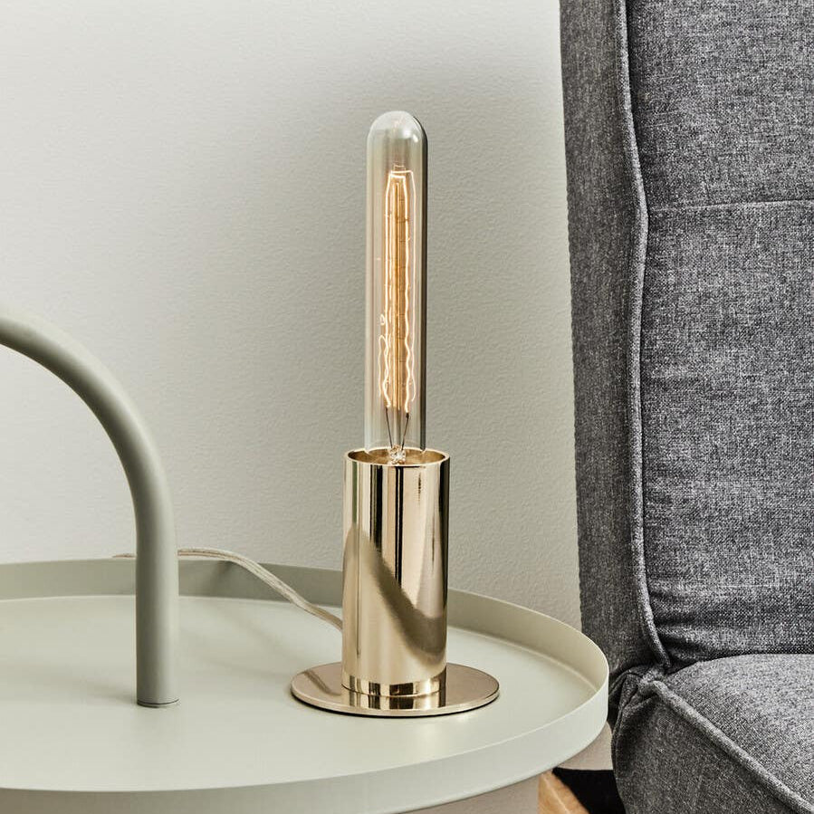 A stylish grey hanging top side table featuring a round surface with a light-colored finish, accompanied by a modern gold lamp with a cylindrical shade, positioned next to a grey upholstered sofa