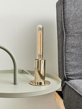 A stylish grey hanging top side table featuring a round surface with a light-colored finish, accompanied by a modern gold lamp with a cylindrical shade, positioned next to a grey upholstered sofa
