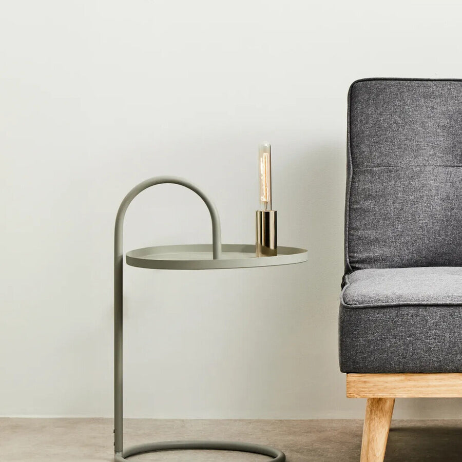 Grey hanging top side table with a circular base and a curved metal frame, positioned next to a modern grey sofa. A small candle holder is placed on the tabletop, which has a sleek, minimalist design. The background features a light-colored wall and a neutral floor
