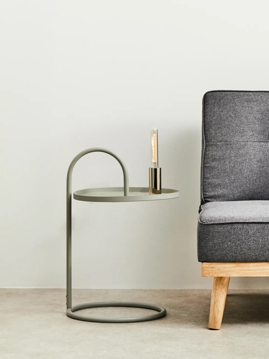 Grey hanging top side table with a circular base and a curved metal frame, positioned next to a modern grey sofa. A small candle holder is placed on the tabletop, which has a sleek, minimalist design. The background features a light-colored wall and a neutral floor
