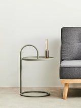 Grey hanging top side table with a circular base and a curved metal frame, positioned next to a modern grey sofa. A small candle holder is placed on the tabletop, which has a sleek, minimalist design. The background features a light-colored wall and a neutral floor