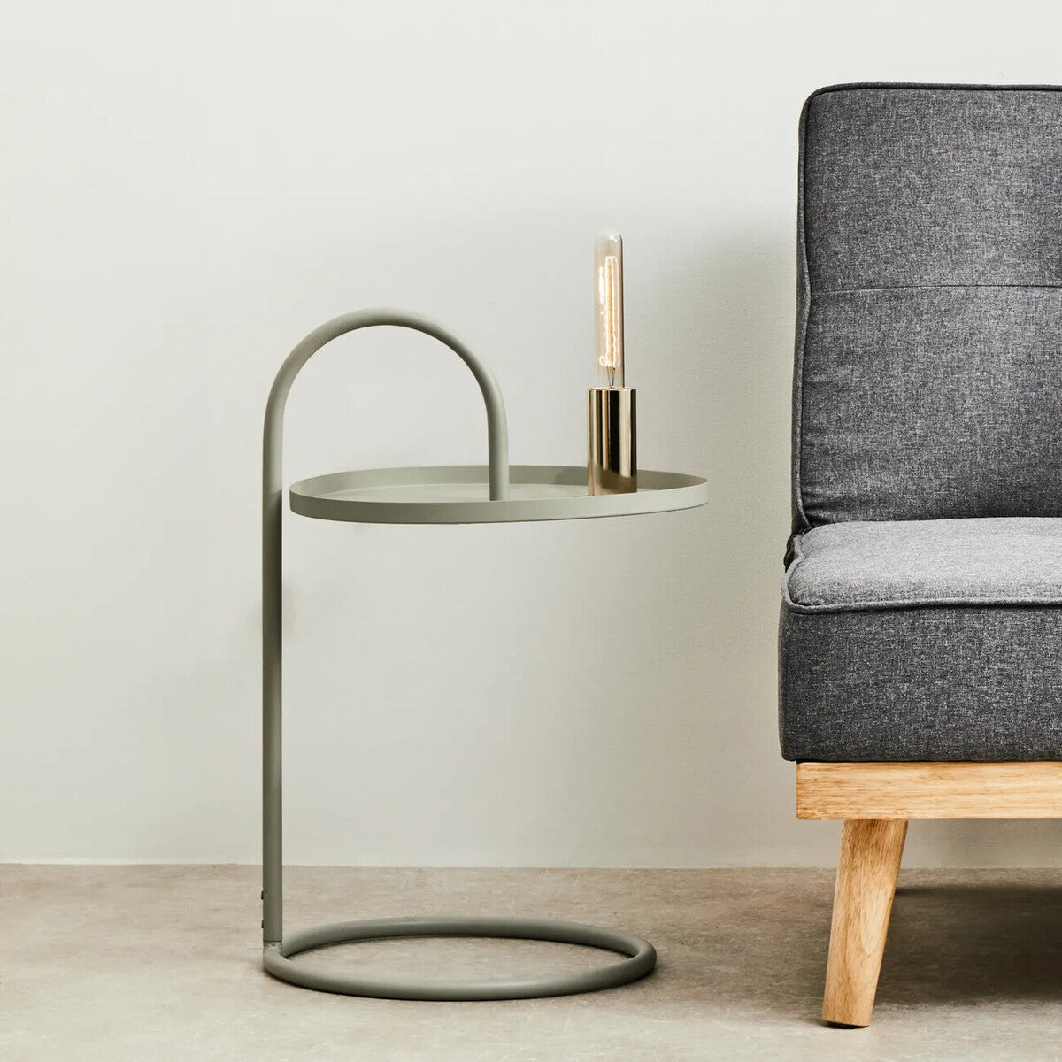 Grey hanging top side table with a circular glass top and a curved metal base, positioned next to a modern grey upholstered sofa with wooden legs. A small candle holder is placed on the table. The background features a neutral wall and floor
