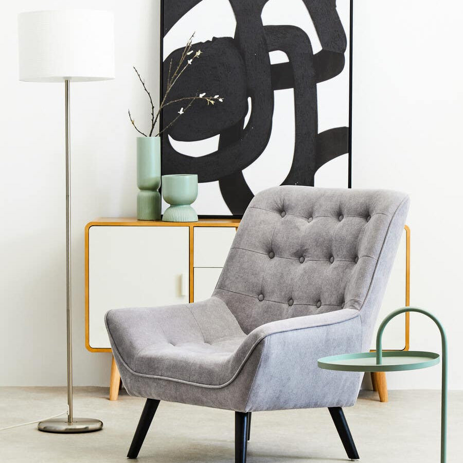 A green hanging top side table with a sleek, modern design featuring a circular base and a curved top, positioned next to a gray upholstered armchair with button detailing. The background includes a minimalist floor lamp and a decorative wall art piece with abstract black lines on a white canvas