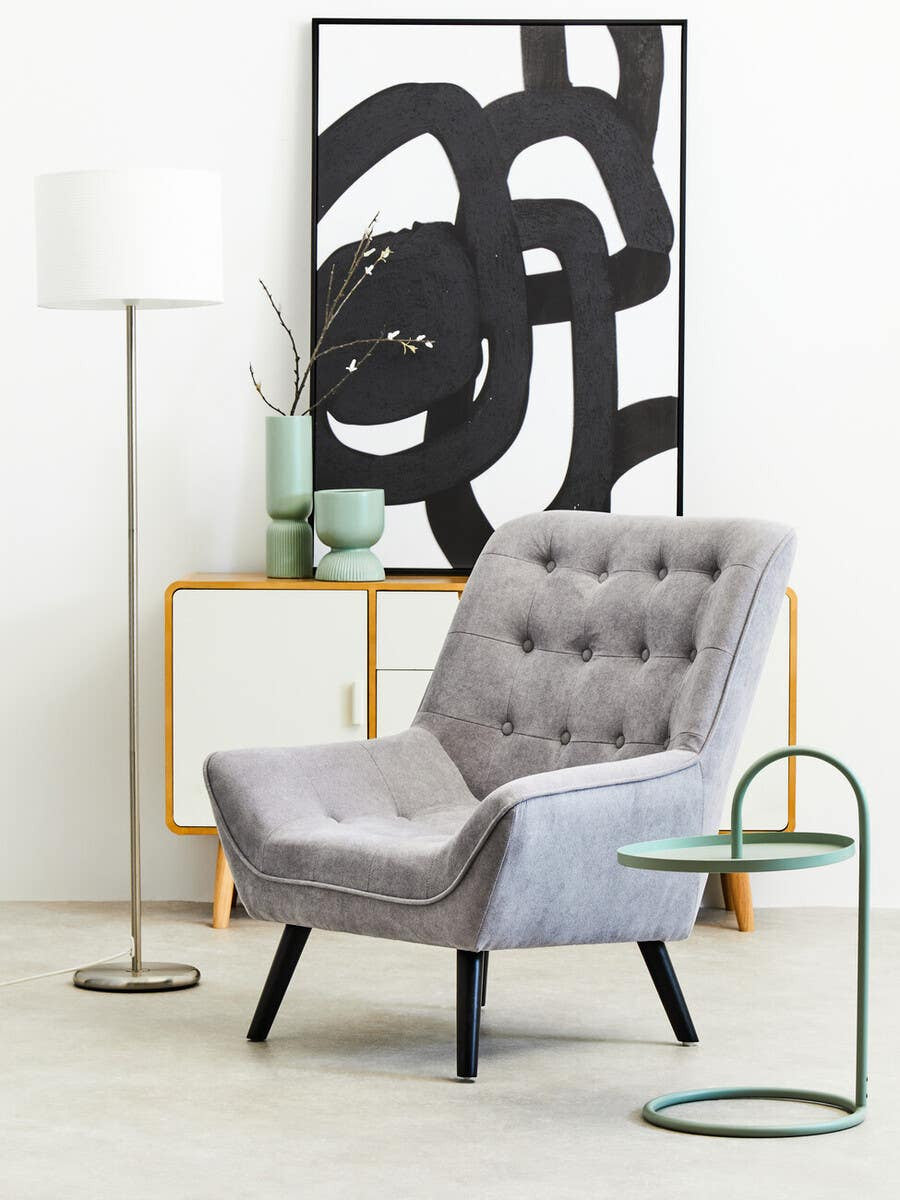 A green hanging top side table with a sleek, modern design featuring a circular base and a curved top, positioned next to a gray upholstered armchair with button detailing. The background includes a minimalist floor lamp and a decorative wall art piece with abstract black lines on a white canvas