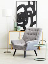 A green hanging top side table with a sleek, modern design featuring a circular base and a curved top, positioned next to a gray upholstered armchair with button detailing. The background includes a minimalist floor lamp and a decorative wall art piece with abstract black lines on a white canvas