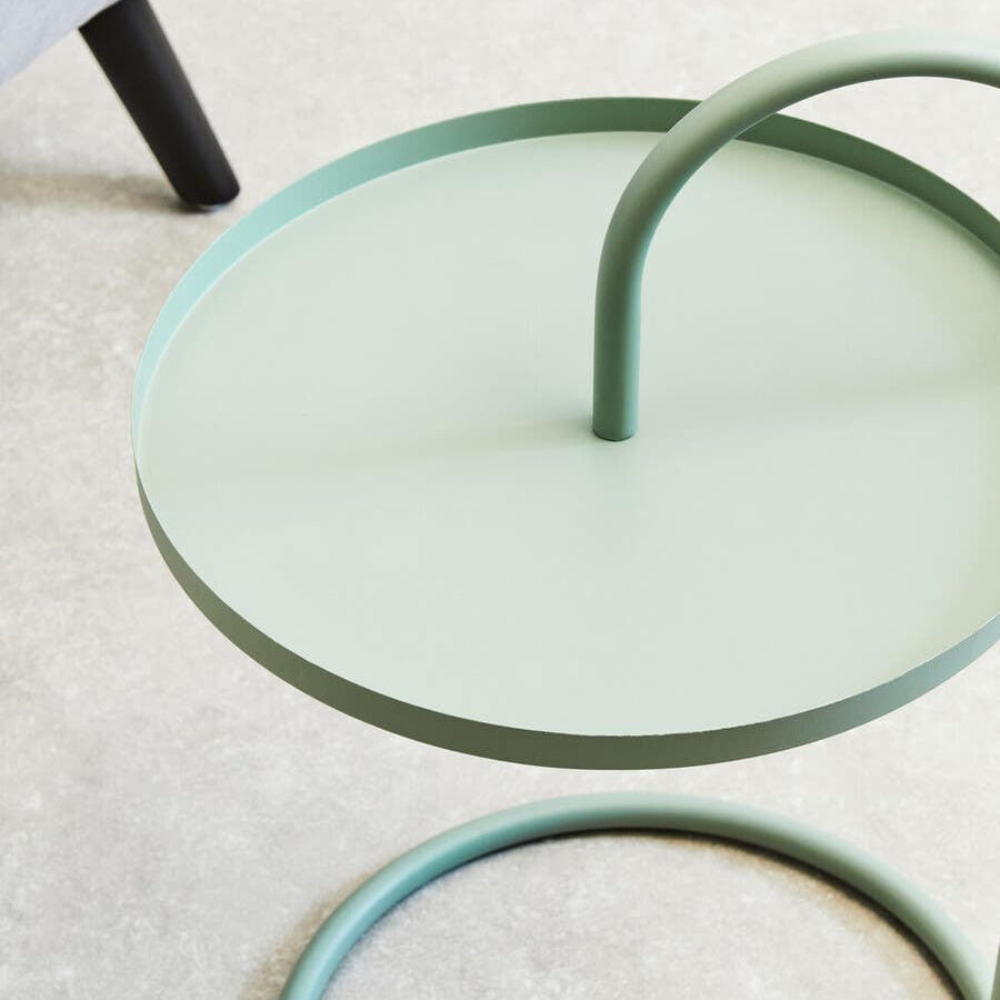 A green hanging top side table with a circular surface and a curved handle, featuring a minimalist design. The table is placed on a light-colored floor, with part of a black leg from another piece of furniture visible in the background