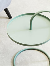 A green hanging top side table with a circular surface and a curved handle, featuring a minimalist design. The table is placed on a light-colored floor, with part of a black leg from another piece of furniture visible in the background