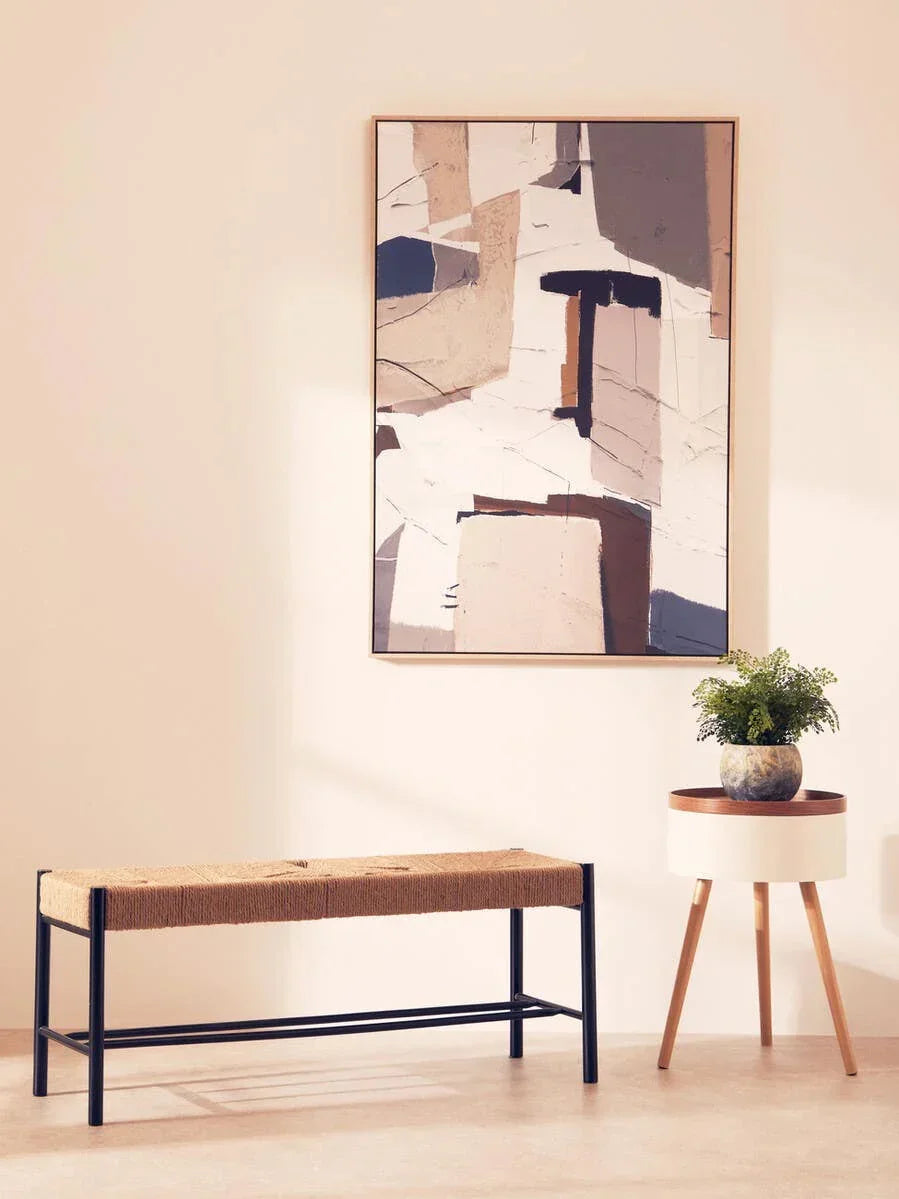 Woven bench with black metal frame beside round wooden side table and abstract wall art