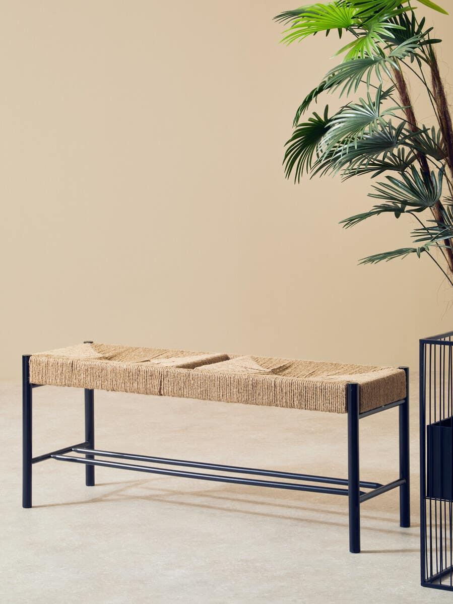 Woven bench with black metal frame beside indoor plant on beige background