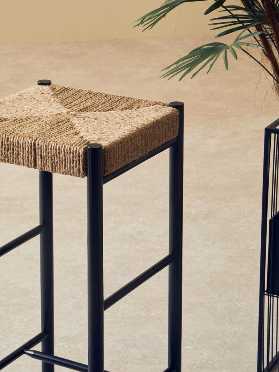 Square bar stool with natural woven seat and black metal frame beside potted plant