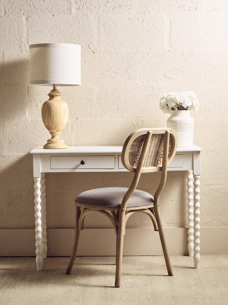 White wooden desk with spindle legs, table lamp, rattan chair and vase of flowers