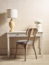 White wooden desk with spindle legs, table lamp, rattan chair and vase of flowers