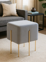 Square grey velvet stool with gold metal legs, placed on a light-colored rug in a cozy living room setting, with a sofa and decorative pillows in the background