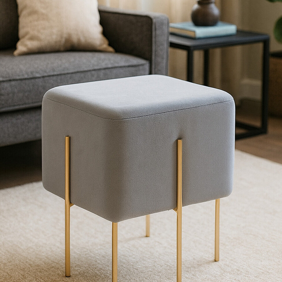 Square grey velvet stool with gold metal legs, placed on a light-colored rug in a cozy living room setting. A sofa and decorative pillow are visible in the background, along with a small table and a plant