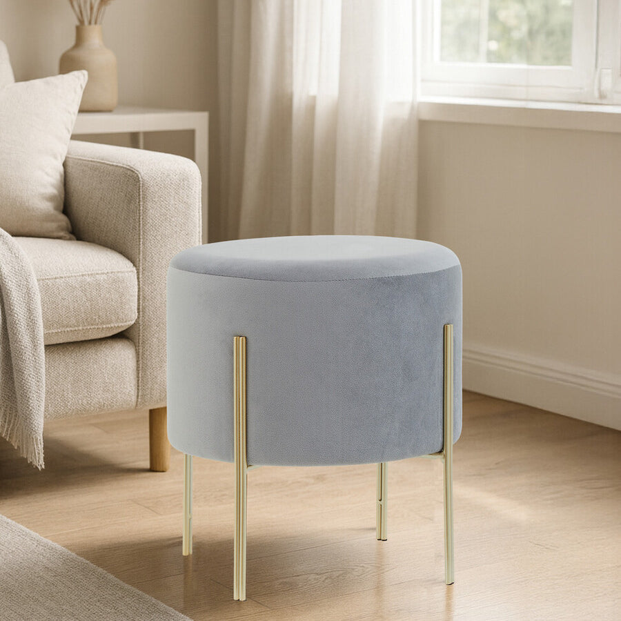 Round grey velvet stool with gold metal legs, placed beside a light-colored sofa in a bright room with natural light streaming through a window. The floor is wooden, and there are soft furnishings in the background