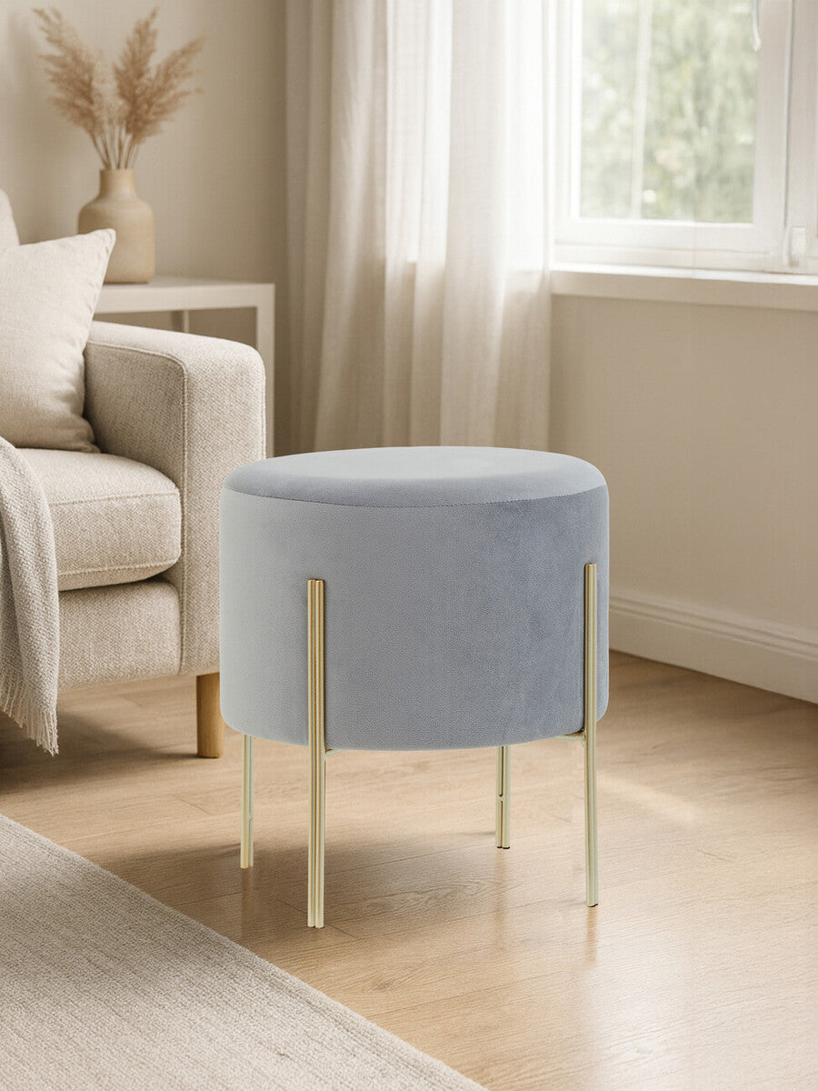 Round grey velvet stool with gold metal legs, placed beside a light-colored sofa in a bright room with natural light streaming through a window. The floor is wooden, and there are soft furnishings in the background