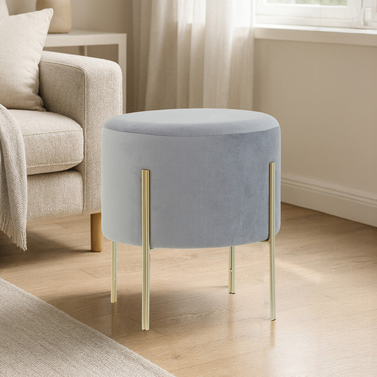 Round grey velvet stool with gold metal legs, placed on a light wooden floor in a well-lit room with a soft beige couch and a window in the background