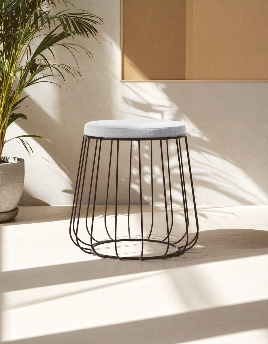 A grey velvet stool with a plush top and a sleek black wire frame, positioned on a light-colored floor with soft shadows and a warm, inviting atmosphere. In the background, there are hints of a cozy living space, including a light-colored sofa and a potted plant