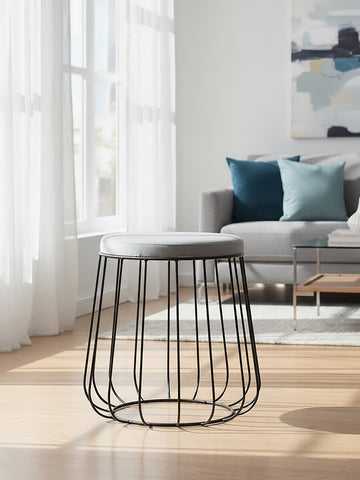 Grey velvet stool with a black metal frame, featuring a rounded top and an open wire design for the base. The stool is positioned on a light wooden floor, with a bright room in the background that includes a sofa and decorative pillows. Natural light filters through large windows, enhancing the modern aesthetic of the space