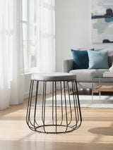 Grey velvet stool with a black metal frame, featuring a rounded top and an open wire design for the base. The stool is positioned on a light wooden floor, with a bright room in the background that includes a sofa and decorative pillows. Natural light filters through large windows, enhancing the modern aesthetic of the space