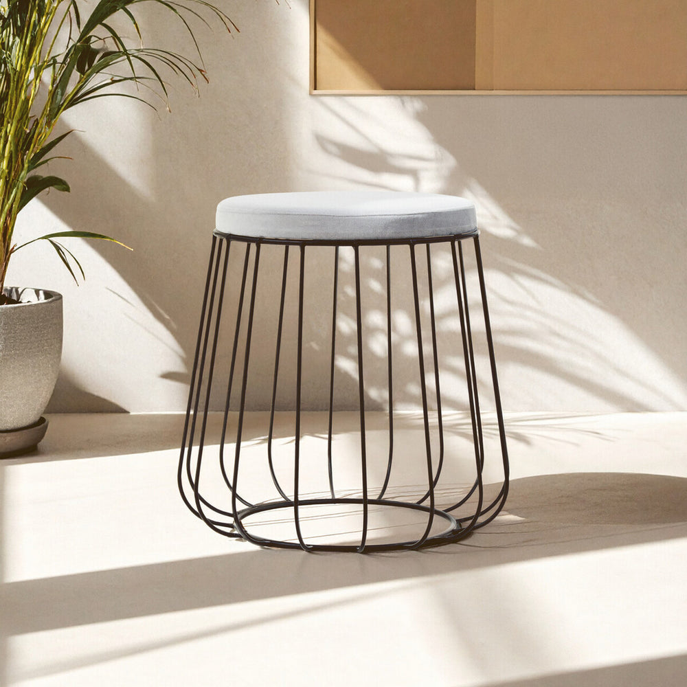 Grey velvet stool with a black wire frame, featuring a round top and an open, airy design. The stool is positioned on a light-colored surface, with soft shadows and natural light illuminating the scene. A potted plant is visible in the background, adding a touch of greenery