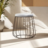 Grey velvet stool with a black wire frame, featuring a round top and an open, airy design. The stool is positioned on a light-colored surface, with soft shadows and natural light illuminating the scene. A potted plant is visible in the background, adding a touch of greenery