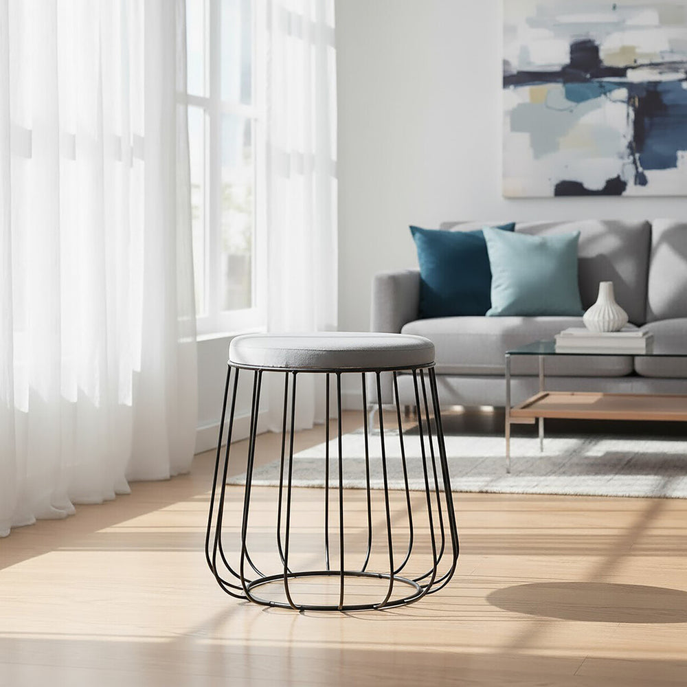 Grey velvet stool with a sleek black metal frame, positioned in a bright room with large windows. The stool features a soft, plush top and a modern, open design. In the background, a comfortable grey sofa with colorful throw pillows and a wooden coffee table are visible, along with light-colored flooring