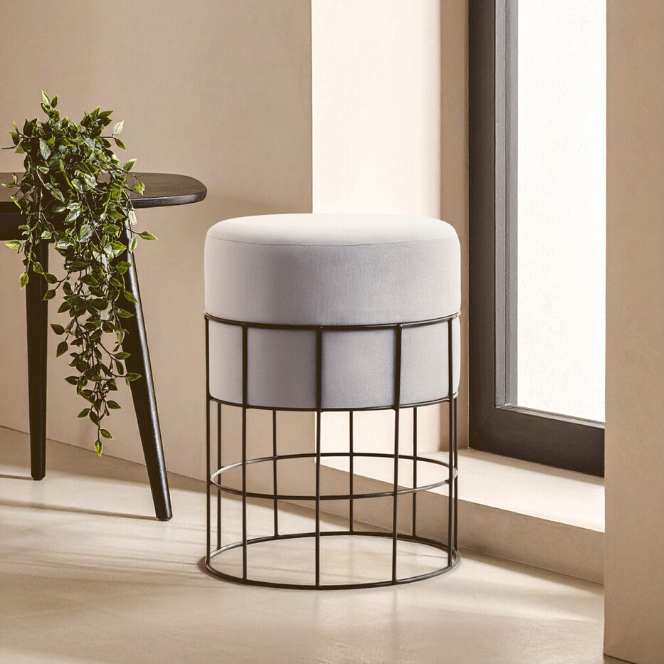 Velvet stool with a round top in light gray, supported by a black metal cage frame, positioned near a window with natural light illuminating the scene. A small plant is visible on a nearby table