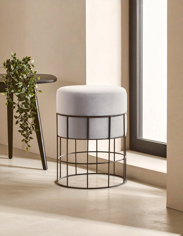 Velvet stool with a round top in light gray, supported by a black metal cage frame, positioned near a window with natural light illuminating the scene. A small plant is visible on a nearby table