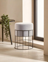 Velvet stool with a round top in light gray, supported by a black metal cage frame, positioned near a window with natural light illuminating the scene. A small plant is visible on a nearby table