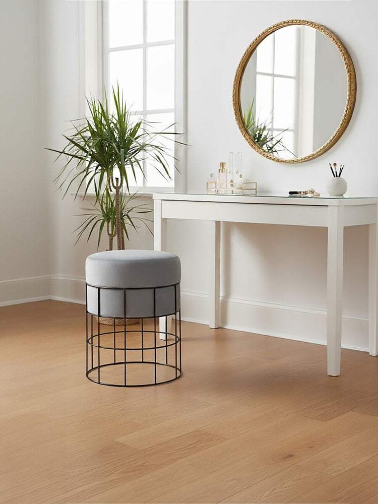 A velvet stool with a gray cushion and a sleek metal cage frame sits on a light wooden floor next to a white console table. A round mirror with a gold frame hangs on the wall above the table, and a potted plant adds a touch of greenery to the scene. Natural light streams in through a window, creating a bright and inviting atmosphere