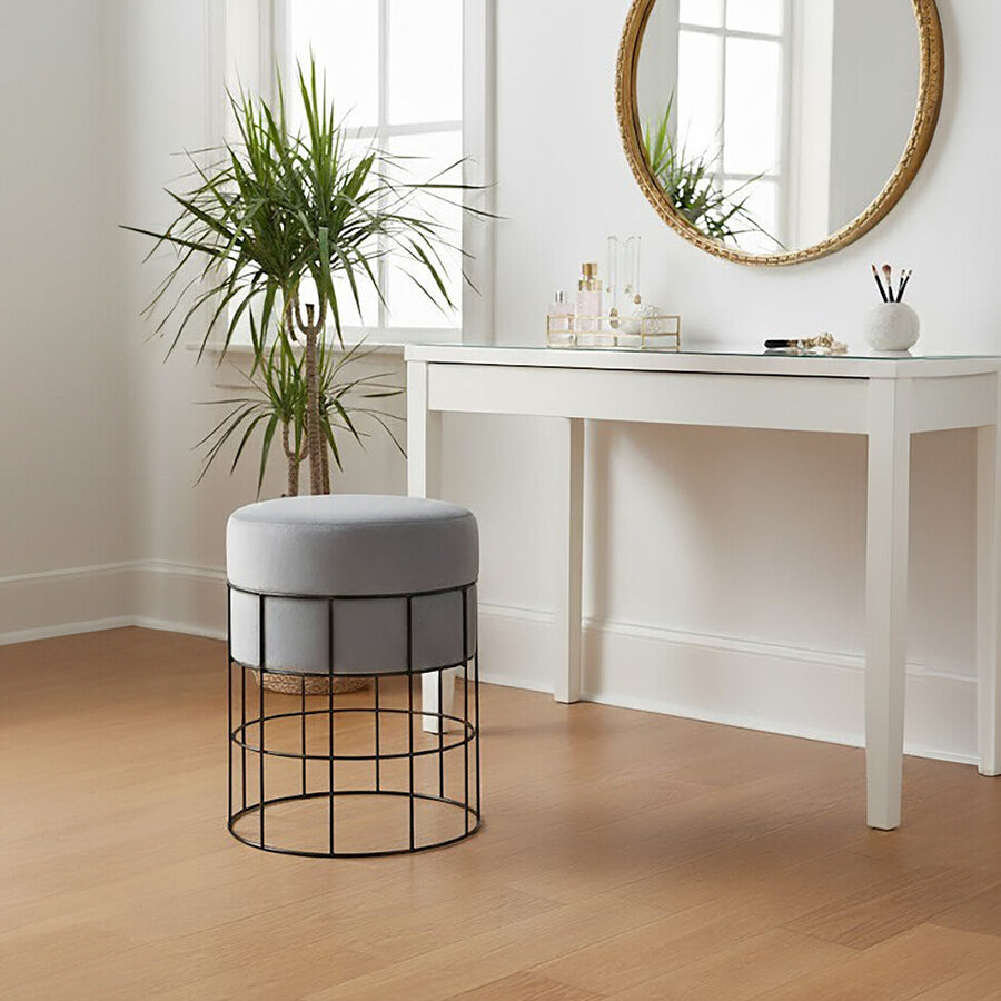 A velvet stool with a gray cushion and a sleek metal cage frame sits on a light wooden floor next to a white console table. A round mirror with a gold frame hangs on the wall above the table, and a potted plant adds a touch of greenery to the scene. Natural light streams in through a window, creating a bright and inviting atmosphere